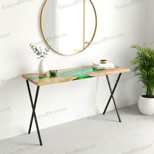 May include: A modern console table with a live edge wooden top and a vibrant green epoxy resin river. The table has black metal legs and is displayed in a well-lit room with a gold-framed mirror and a potted plant.