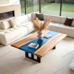 May include: A modern coffee table with a live edge wood design and a blue epoxy resin center. The table has a rectangular shape, supported by black metal legs. The table is surrounded by a white sofa and a neutral-colored rug.