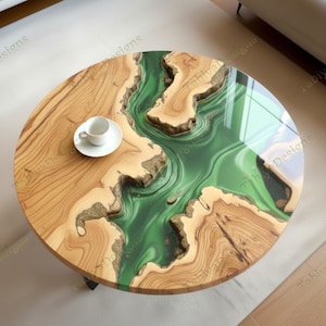 Op de afbeelding: Ronde houten salontafel met een rivier van groene epoxyhars. De tafel heeft een natuurlijk houtnerfpatroon en een glanzende afwerking. Een witte kop en schotel staan op het houten oppervlak.