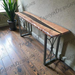 Op de afbeelding: Een houten console met een levende rand en een donkere epoxyharsinleg. De tafel heeft een rechthoekig blad en wordt ondersteund door zwarte metalen poten met decoratieve krullen. De tafel is ongeveer 152 cm lang.