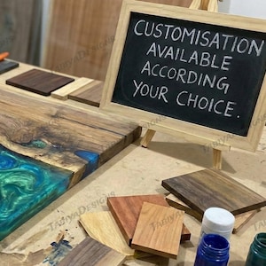 May include: A wooden table with a blue and green epoxy resin river. A small chalkboard on an easel reads "CUSTOMISATION AVAILABLE ACCORDING YOUR CHOICE." Various wood samples and paint bottles are also visible.