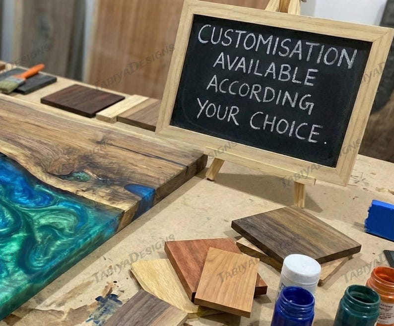 May include: A wooden table with a blue and green epoxy resin river. A small chalkboard on an easel reads "CUSTOMISATION AVAILABLE ACCORDING YOUR CHOICE." Various wood samples and paint bottles are also visible.