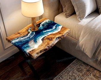 May include: A handcrafted side table with a live edge wooden top. The table features a blue and white resin design resembling ocean waves. A gold-toned lamp with a beige shade sits on the table. The table is supported by black metal legs.