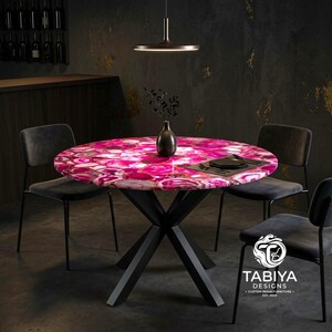 May include: A round dining table featuring a vibrant pink agate stone top with a polished finish, supported by a black metal X-shaped base. Three dark gray upholstered chairs surround the table. A black vase sits in the center of the table.