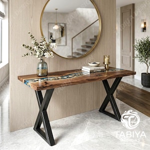 Peut inclure: Une console rectangulaire dotée d'un plateau en bois naturel avec une incrustation centrale en résine bleue contenant des pierres. La table est soutenue par deux pieds en métal noir en forme de X. Un vase en verre avec des plantes est posé.