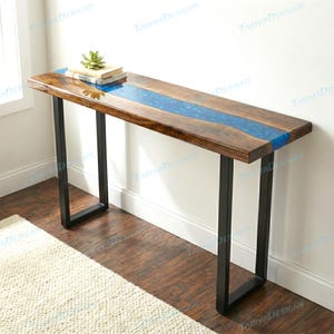 May include: A wooden console table with a blue resin river design. The tabletop is made of dark wood and features a blue resin inlay. The table has black metal legs and is decorated with a small succulent and books.