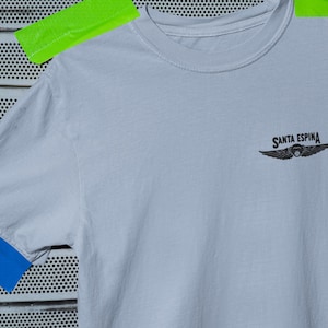 Op de afbeelding: Een lichtblauw t-shirt met de tekst "SANTA ESPINA" en een gevleugeld embleem. Het shirt heeft een ronde hals en korte mouwen. Het shirt is met groen en blauw plakband op een metalen oppervlak geplakt.