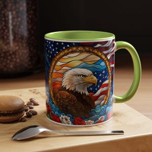 May include: A ceramic mug with a patriotic design featuring a bald eagle, American flag, and floral elements. The mug has a green interior and handle. The design is set against a white background.