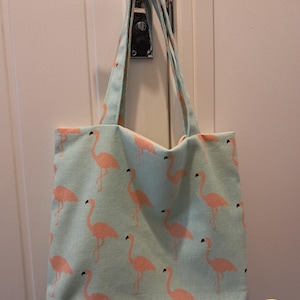 May include: A pale blue tote bag featuring a pattern of pink flamingos. The bag has two handles and is suspended from a door handle. The bag is made of fabric and is rectangular in shape.