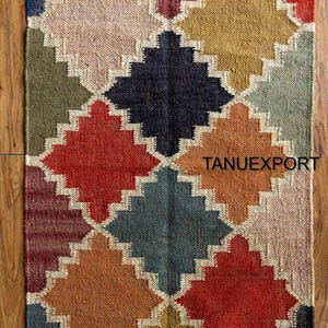 May include: A rectangular rug with a geometric diamond pattern in various colors, including red, blue, yellow, and beige. The rug has a fringed edge and the text "TANUEXPORT".