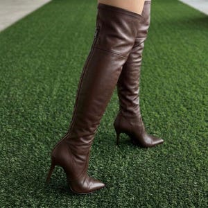 K&ouml;nnte beinhalten: Ein Paar dunkelbraune Overknee-Stiefel mit glatter Lederoptik, spitzer Zehenpartie und schmalem Stiletto-Absatz. Die Stiefel verf&uuml;gen &uuml;ber einen durchgehenden vertikalen Rei&szlig;verschluss an der Au&szlig;enseite des Schafts, der bis zum oberen Rand reicht.