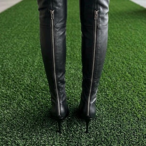 K&ouml;nnte beinhalten: Ein Paar schwarze Overknee-Stiefel mit einem durchgehenden, silberfarbenen Metallrei&szlig;verschluss an der R&uuml;ckseite jedes Schafts, der in einem schmalen Stiletto-Absatz endet. Die Stiefel bestehen aus einem glatten, mattschwarzen Material.