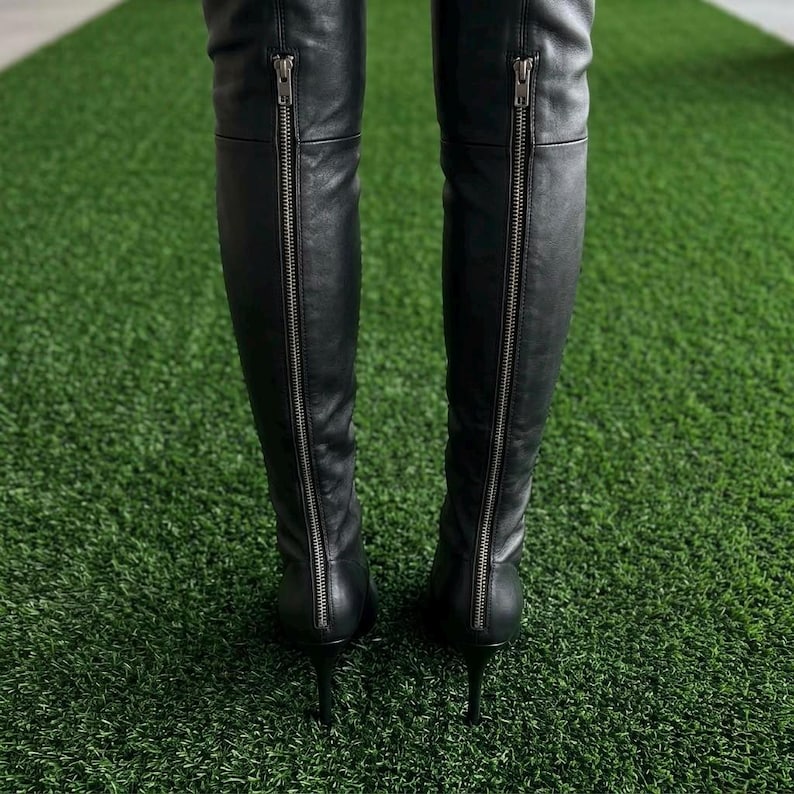 K&ouml;nnte beinhalten: Ein Paar schwarze Overknee-Stiefel mit einem durchgehenden, silberfarbenen Metallrei&szlig;verschluss an der R&uuml;ckseite jedes Schafts, der in einem schmalen Stiletto-Absatz endet. Die Stiefel bestehen aus einem glatten, mattschwarzen Material.