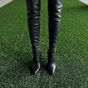 K&ouml;nnte beinhalten: Ein Paar schwarze Overknee-Stiefel mit glatter Oberfl&auml;che, spitzer Zehenpartie und einer vertikalen Naht entlang des Schafts. Die Stiefel reichen &uuml;ber das Knie, haben eine eng anliegende Passform und sind mit einem Stiletto-Absatz versehen.