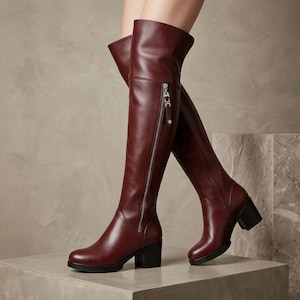 Könnte beinhalten: Ein Paar dunkelburgunderrote Overknee-Stiefel mit glatter Oberfläche, abgerundeter Zehenpartie und Blockabsatz. Jeder Stiefel verfügt über einen durchgehenden silberfarbenen seitlichen Reißverschluss mit Marken-Zipper vom Knöchel bis zum Schaft.