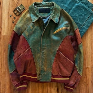 Puede incluir: Chaqueta de cuero vintage en tonos verdes, marrones y burdeos. La chaqueta presenta un cuello, un bolsillo con cremallera y un dobladillo acanalado con rayas amarillas. Un cinturón a juego y un trozo de tela están junto a la chaqueta.