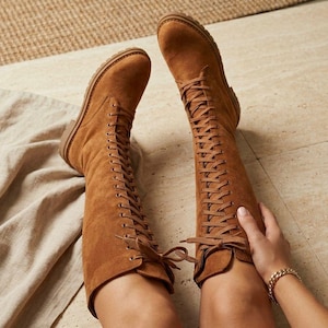 May include: A pair of knee-high tan suede boots featuring a full-length lace-up front with matching tan laces, rounded toes, and a flat, textured rubber sole. The boots extend to the knee and are tied at the top with a bow.