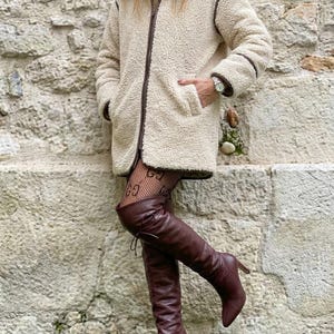 Può includere: Una giacca in pile sherpa testurizzato color crema con cerniera frontale, colletto ripiegato e finiture marrone scuro lungo le cuciture e la cerniera. È abbinata a stivali alti in pelle marrone con tacco.