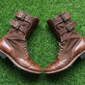 May include: A pair of mid-calf boots crafted from brown leather and dark brown fabric. These boots feature a lace-up front closure, two adjustable leather straps with dark metal buckles across the upper shaft, and a thick, dark lugged rubber sole.