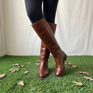 May include: A pair of knee-high brown leather boots with a smooth finish, rounded toes, and a low block heel. The boots feature a simple, unadorned shaft design that reaches just below the knee, providing a clean and classic silhouette for footwear.