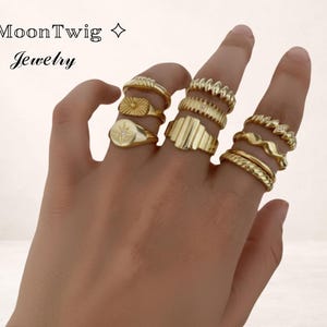 Peut inclure: Une main présentant une collection de bagues dorées. Les bagues présentent divers motifs, dont un soleil, une étoile, des textures côtelées et des motifs ondulés. Les bagues proviennent de la collection MoonTwig Jewelry.