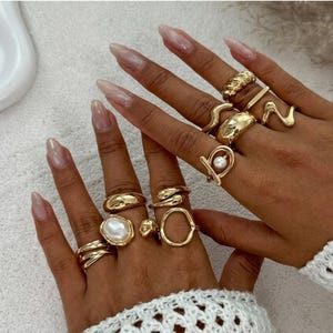 Puede incluir: Una colección de anillos dorados en varios diseños, incluyendo un anillo con perla, un anillo retorcido y un anillo con forma geométrica. Los anillos se muestran en una mano con uñas largas y pulidas, mostrando los detalles de la joyería.