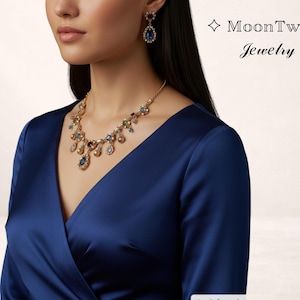Puede incluir: Un collar dorado y pendientes a juego con piedras preciosas de colores y una gran piedra azul en forma de lágrima. Las joyas se muestran sobre una persona que lleva un vestido de satén azul oscuro. El texto "MoonTwig Jewelry" está en la esquina superior derecha.