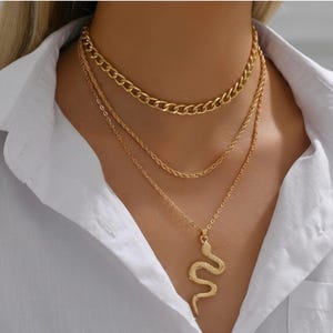 May include: A gold-colored layered necklace set. The top layer is a thick chain, the middle is a rope chain, and the bottom features a snake pendant. The necklace is worn over a white collared shirt.