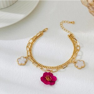 Puede incluir: Pulsera dorada de doble capa con abalorios de flores y tréboles. La pulsera presenta un vibrante abalorio de flor fucsia y dos abalorios de trébol blanco. Es una joya delicada.