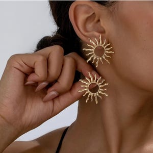Może przedstawiać: Złote kolczyki w kształcie słońca. Każdy kolczyk ma centralny okrągły wzór z promieniami. Kolczyki są pokazane na uchu i trzymane przez dłoń, prezentując ich design.