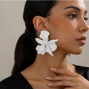 Peut inclure: Boucles d'oreilles blanches en forme de fleur. Les boucles d'oreilles sont grandes et présentent un design de pétales détaillé. Elles sont portées par une personne aux cheveux foncés et un haut noir. L'arrière-plan est de couleur neutre.
