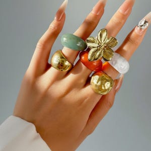 Könnte beinhalten: Eine Hand, die eine Sammlung von Ringen in verschiedenen Stilen und Farben trägt. Die Ringe umfassen eine goldene Blume, eine goldene Kugel, ein grünes Oval, ein orangefarbenes Oval und einen klaren, marmorierten Ring.