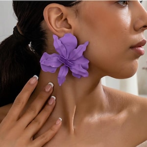 Peut inclure: Gros plan d'une personne portant une grande boucle d'oreille en forme de fleur violette. La boucle d'oreille est une réplique détaillée d'une fleur avec de multiples pétales. La peau de la personne est claire et ses ongles sont manucurés à la française. L'arrière-plan est flou.