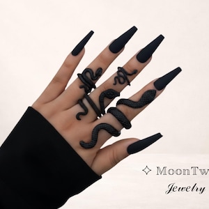 Könnte beinhalten: Eine Hand mit mehreren schwarzen Schlangenringen. Die Ringe sind in verschiedenen Designs um die Finger gewickelt. Die Nägel sind lang und schwarz lackiert. Der Text "MoonTwig Jewelry" ist ebenfalls enthalten.