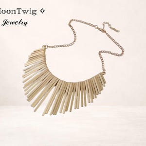 Puede incluir: Un collar dorado con diseño de flecos. El collar presenta una delicada cadena y un colgante compuesto por numerosos trozos rectangulares finos dispuestos en forma curva. El texto "MoonTwig Jewelry" está en la esquina superior izquierda.
