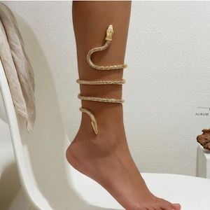 Könnte beinhalten: Goldfarbenes Schlangen-Armband und Fußkettchen. Der Schmuck wickelt sich um Bein und Fuß und schafft ein stilvolles Accessoire. Kopf und Schwanz der Schlange sind sichtbar und fügen Details hinzu. Der Schmuck ist wahrscheinlich aus Metall.