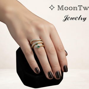 Könnte beinhalten: Goldfarbener Schlangenring mit funkelnden klaren und grünen Edelsteinen. Der Ring wickelt sich spiralförmig um den Finger. Der Ring wird auf einer schwarzen Samtbox präsentiert. Der Text "MoonTwig Jewelry" befindet sich oben rechts.