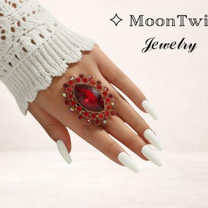 Könnte beinhalten: Ein auffälliger Ring mit einem ovalen, roten Mittelstein, umgeben von kleineren roten und klaren Kristallen. Der Ring wird auf einer Hand mit langen, weißen Nägeln präsentiert. Der Text "MoonTwig Jewelry" ist sichtbar.