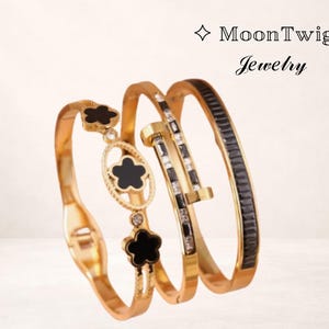 May include: Gold-toned bracelets with black and clear accents. One bracelet features a clover design, another has a nail head design, and the third has black rectangular stones. The MoonTwig Jewelry logo is in the top right corner.