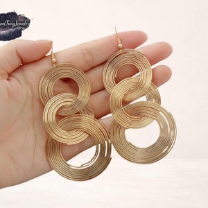 Puede incluir: Pendientes de aro dorados y enlazados. Los pendientes presentan múltiples aros circulares de diferentes tamaños, creando un efecto colgante en capas. El diseño es moderno y geométrico, adecuado para el uso diario o ocasiones especiales. Los pendientes se sostienen en una mano.