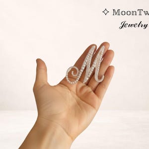 Może przedstawiać: Srebrna litera "M" inkrustowana małymi, przezroczystymi kryształami i perłami. Biżuteria trzymana jest w dłoni na białym tle. Nazwa marki "MoonTwig Jewelry" jest wyświetlana nad literą.