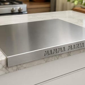 Op de afbeelding: Een rechthoekige fornuisafdekplaat van roestvrij staal met een gladde, geborstelde metalen afwerking. De voorste verticale rand is voorzien van de lasergesneden tekst MAMMA MARIA in een strak, schreefloos lettertype voor in de keuken.