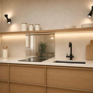Può includere: Una cucina con armadi in legno chiaro, un piano di lavoro bianco e un rubinetto nero. Due applique nere illuminano una mensola in legno con barattoli e piatti bianchi. Un piano cottura e un lavello neri sono integrati nel piano di lavoro.