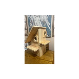 May include: A wooden birdhouse with a pitched roof and two circular entry holes. The birdhouse has two small perches and is made of light-colored wood. The design is simple and functional, suitable for attracting birds to a garden or yard.