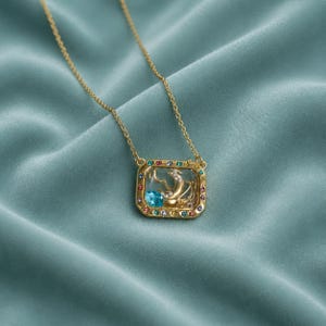 May include: A gold-toned necklace featuring a rectangular pendant with a clear center. The pendant frame is set with small, multi-colored gemstones. Inside the clear center is a gold crescent moon shape and a single, bright blue oval gemstone.