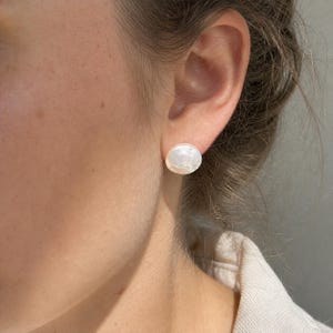 Könnte beinhalten: Ein einzelner runder, weißer, schillernder Perlenstecker mit einer glatten, glänzenden Oberfläche. Der Ohrring ist am Ohrläppchen befestigt und zeigt eine klassische, kreisförmige Form sowie ein reflektierendes, perlmuttartiges Finish, das das Licht einfängt.