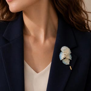 May include: A lapel pin featuring a cluster of three fan-shaped, textured elements in white with blue-tipped edges. The decorative piece is attached to a gold-toned metal pin stem, secured to the fabric of a dark navy blue blazer.