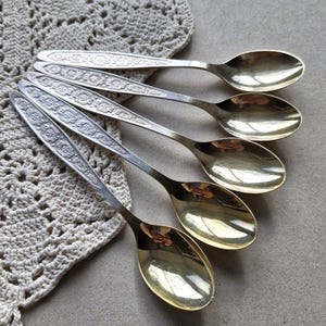 Op de afbeelding: Een set van vier goudkleurige metalen lepels in een waaiervormige opstelling. Elke lepel heeft een lange, slanke steel versierd met een herhalend geometrisch reliëfpatroon. De lepels hebben een glad, gepolijst en reflecterend oppervlak.