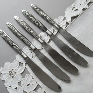 May include: Set of six silver dinner knives with floral designs on the handles. The knives are arranged in a row on a white lace tablecloth.