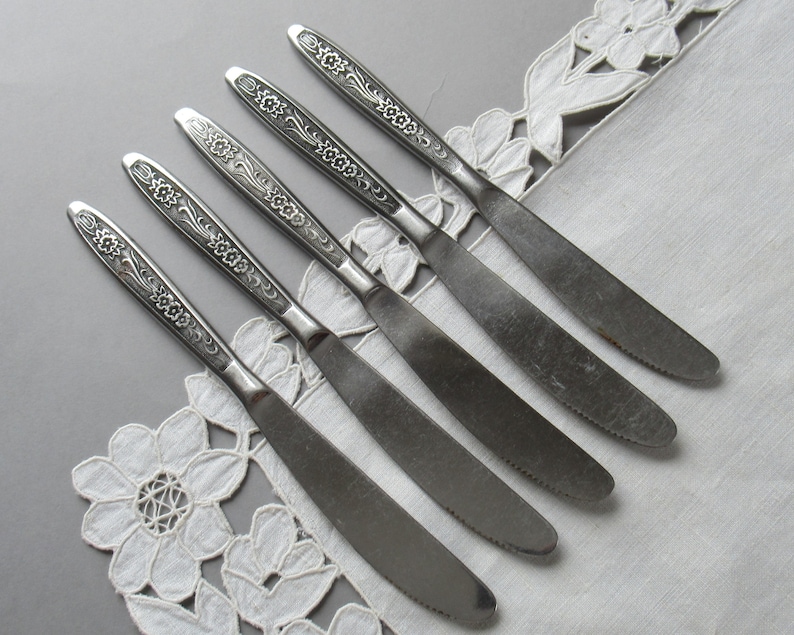 May include: Set of six silver dinner knives with floral designs on the handles. The knives are arranged in a row on a white lace tablecloth.